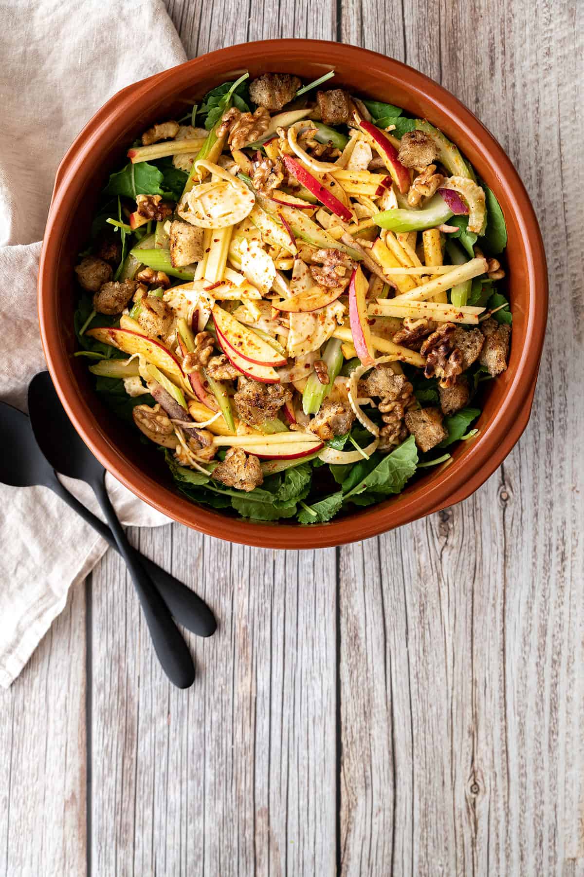 A terracotta dish with apple, fennel and celery salad in it. Pictured alongside a teatowel and serving spoons.