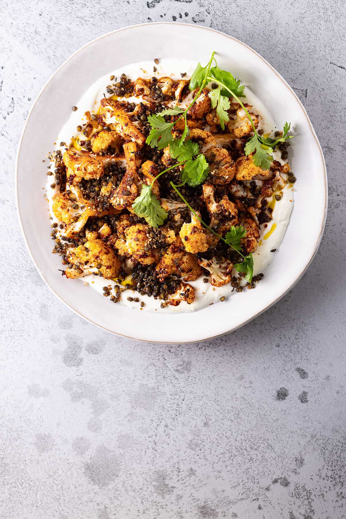 A white plate smeared with yoghurt, topped with cauliflower that has been tossed in harissa paste and roasted, and a scattering of crunchy fried green lentils.