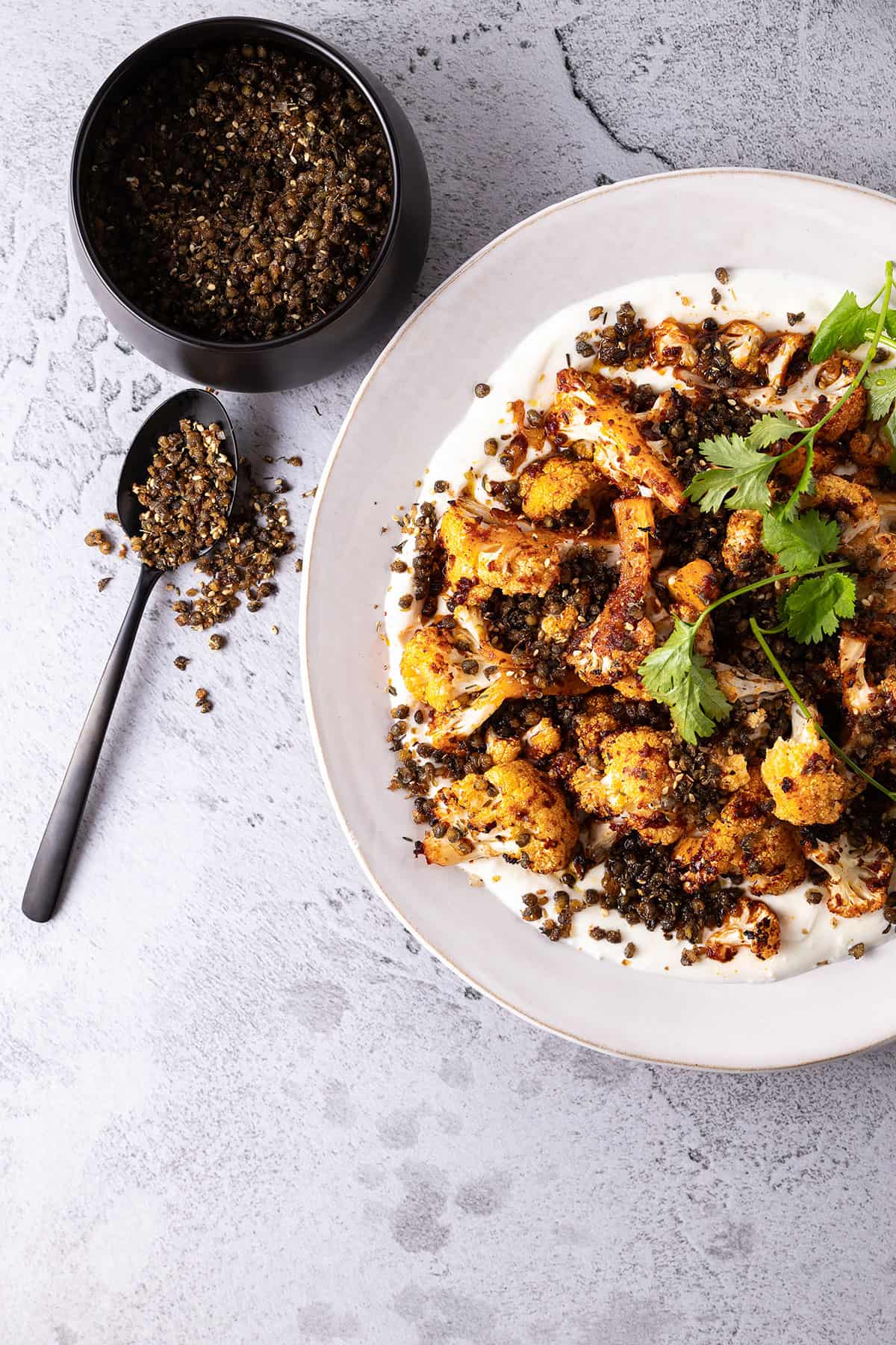 A white plate smeared with yoghurt, topped with cauliflower that has been tossed in harissa paste and roasted, and a scattering of crunchy fried green lentils. Alongside the plate is a bowl of additional fried lentils, a spoon and some spilled lentils. 