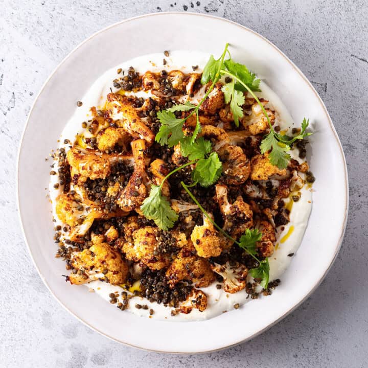 A white plate smeared with yoghurt, topped with cauliflower that has been tossed in harissa paste and roasted, and a scattering of crunchy fried green lentils.n