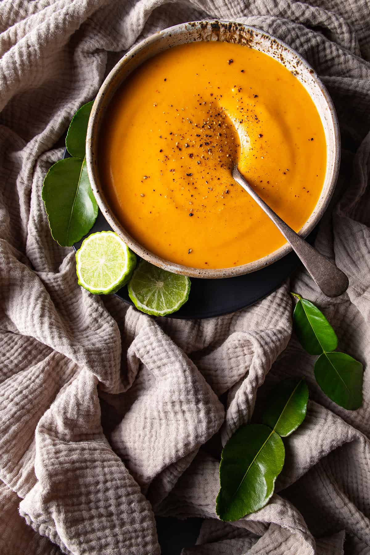 A rustic bowl of bright orange soup, sitting on a beige muslin cloth. Cut makrut limes and makrut lime leaves sitting nearby.