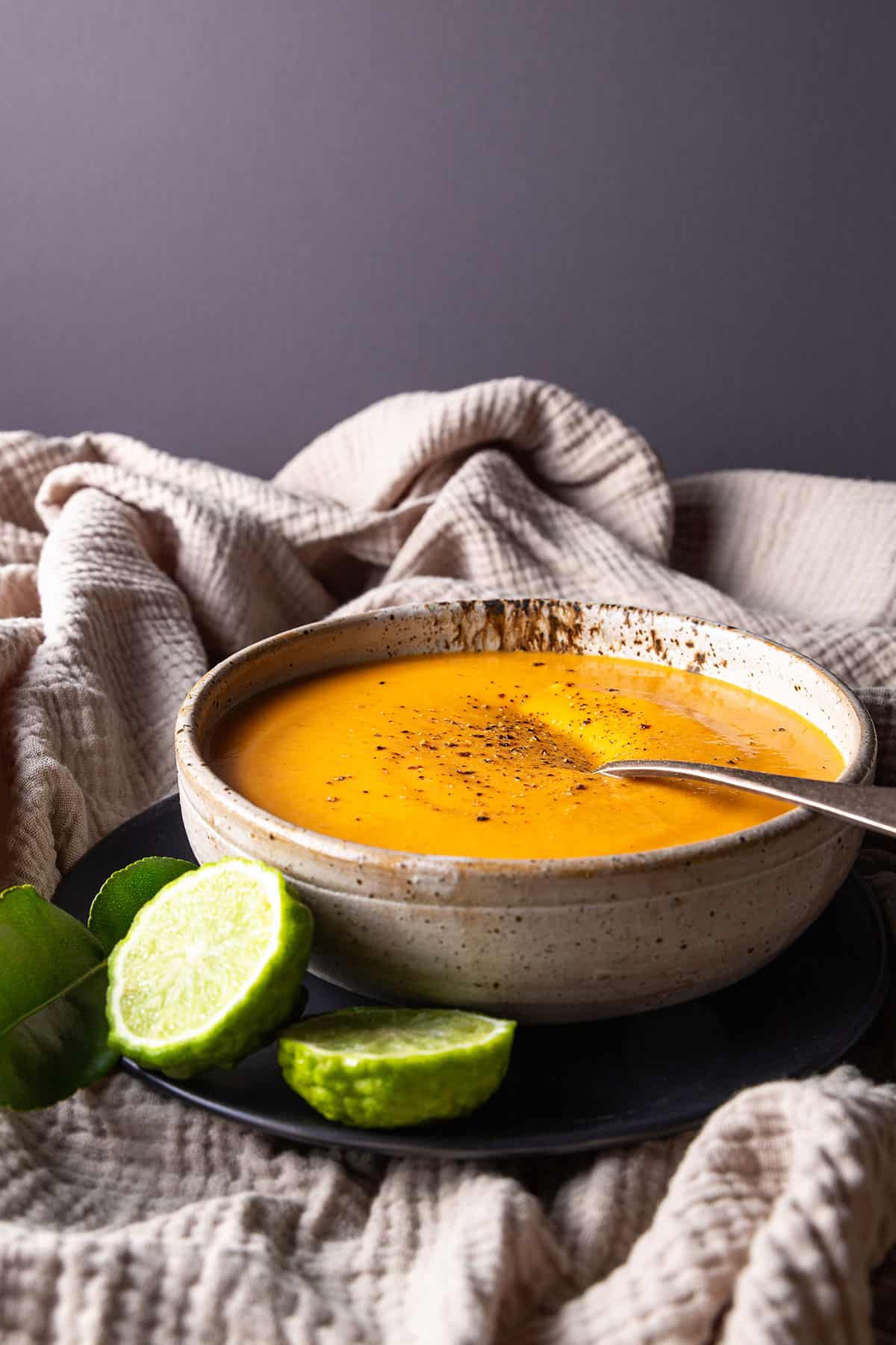 A rustic bowl of bright orange soup, sitting on a beige muslin cloth. Cut makrut limes and makrut lime leaves sitting nearby.