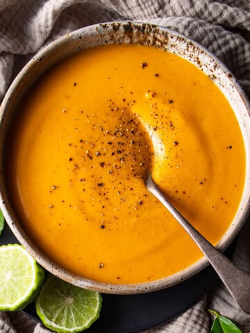 A rustic bowl of bright orange soup, sitting on a beige muslin cloth. Cut makrut limes and makrut lime leaves sitting nearby.