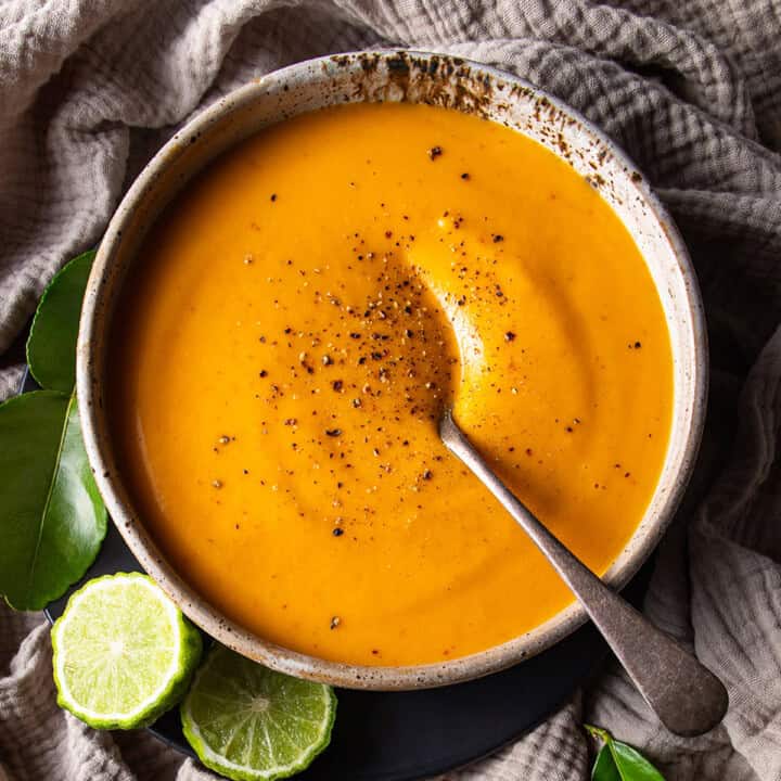 A rustic bowl of bright orange soup, sitting on a beige muslin cloth. Cut makrut limes and makrut lime leaves sitting nearby.