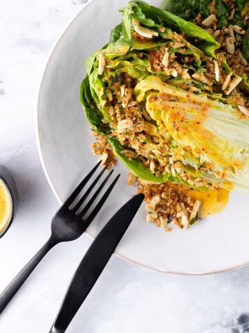 A white plate with half a baby cos lettuce sitting on it, drizzled with bright orange dressing, and scattered with a crunchy topping that includes almonds and breadcrumbs.